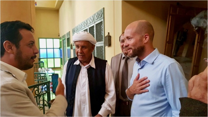 ID: Three men talking, one in blue shirt with hand over heart, another in white shirt and black vest with white turban, another in white shirt with hand raised.